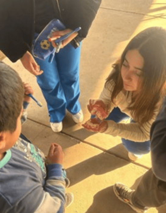 Estudiante de  Odontología en cunclillas explicandole a un niño con un modelo de l boca humana en las manos. Mientras otra estudiante sostiene un estuche con pasta y cepillo de dientes